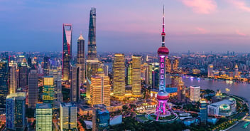 A panorama of the Shanghai skyline can be seen in the twilights hours, the sun having descended upon the horizon as a partially cloudy pink-tinged blue and grey sky succumbs to the onset of dusk, while the city below appears brightly illuminated in varying shades of gold, yellow and red, with many of its iconic buildings standing proudly upright, notably the Oriental Pearl Tower which features prominently to the right of the foreground, glistening in hot pink and purple tones, whilst a cluster of other skyscrapers, including the Shanghai World Financial Center and Shanghai Tower, extend upwards to the left of the frame. 