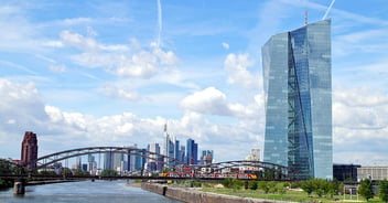 Under a blue and sun-drenched sky, with minimal cumulus cloud cover, the skyscraper building of the European Central Bank is seen standing tall in the foreground, with the Main river remaining visible as it flows into the background, passing under a steel truss bridge, as Frankfurt's skyline appears clustered on the horizon