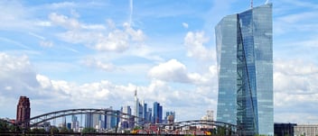 Under a blue and sun-drenched sky, with minimal cumulus cloud cover, the skyscraper building of the European Central Bank is seen standing tall in the foreground, with the Main river remaining visible as it flows into the background, passing under a steel truss bridge, as Frankfurt's skyline appears clustered on the horizon