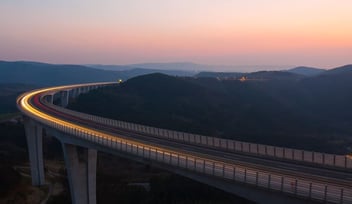 A high bridge in the foreground with a yellow streak of light from the cars. Rolling hills and pink skies in the background. 