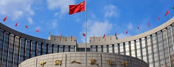 Viewed head on, the concentric cylindrical concrete roof and upper floors of the People's Bank of China building can be seen, centrepieced by a large, billowing Chinese flag, while smaller Chinese flags stand atop the structure in the background.