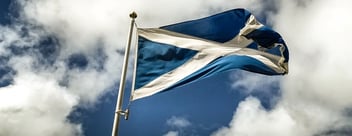 Seen from below, a Scottish flag is seen billowing in the wind atop a flagpole, against a partially cloudy blue sky, exhibiting similar blue and white tones to the flag itself.